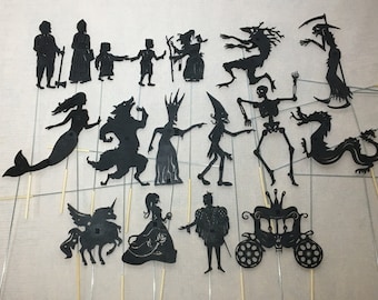May include: A set of 17 black shadow puppets, including a mermaid, a wolf, a unicorn, a princess, a prince, a carriage, a skeleton, a dragon, a witch, a goblin, and more. The puppets are made of black paper and are attached to sticks.