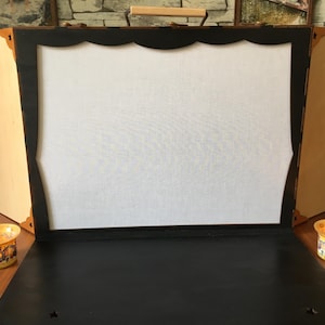 May include: A wooden puppet theater with a white fabric screen and a black frame. The theater has a handle on the top and is closed. Two small candle holders are on the table in front of the theater.