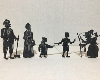 May include: A set of six black silhouette figures, including a man with a staff, a woman with a basket, a girl, a boy, and two witches with broomsticks. The figures are made of paper or cardboard and are designed for shadow puppetry.