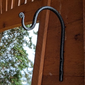 May include: Two black metal hooks are mounted on a wooden wall. The hooks are curved and have a decorative swirl at the top.