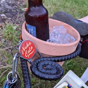 May include: A brown glass bottle of Nuka Cola is in a terracotta flower pot with ice. A black metal bottle holder is attached to the pot. A keychain with a Nuka Cola bottle opener is attached to the holder.