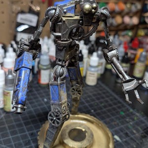 May include: A detailed steampunk robot figurine with a metallic finish and blue accents. The robot features intricate gear and mechanical details, standing on a gold-colored base. The arms end in claw-like hands.