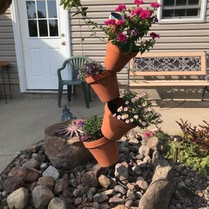 May include: A vertical cascading planter structure made of four stacked terracotta pots angled in alternating directions. The pots are filled with vibrant pink and white flowers and green foliage, supported by a central rod anchored in a rock garden.