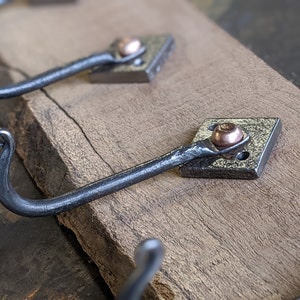 May include: Close-up of three metal hooks with a copper accent on a wooden surface. The hooks are hand-forged and have a rustic, industrial look.