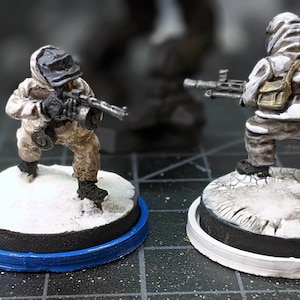 May include: Two painted miniature figures of soldiers wearing winter camouflage gear and holding rifles. The figures are standing on round bases with a snowy, textured surface.