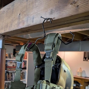 May include: A green tactical vest and a tan helmet hanging on a black metal hook attached to a wooden beam.