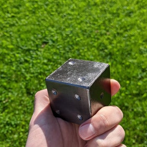 May include: A black metal dice with rounded edges and five dots on the visible side. The dice is being held in a hand against a green grassy background.