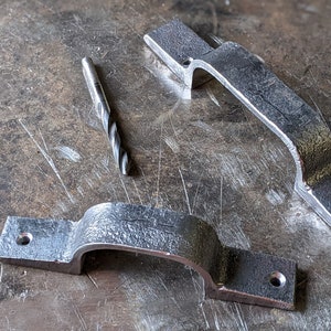 May include: Two silver metal handles with rounded edges and holes for screws. A drill bit is lying on a metal workbench.