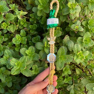 May include: A gold and beige rope charm with various religious and lucky charms, including a green pouch with a dollar bill, an angel, and a saint figure. The charm is held against a backdrop of green leaves.