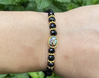 May include: A beaded bracelet featuring faceted black beads and small gold-toned metallic spacers. The center of the bracelet displays an oval-shaped charm with a colorful religious icon set in a gold-toned frame, secured by a black cord closure.