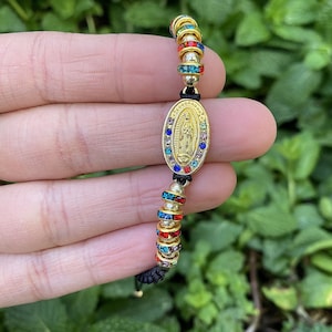 May include: A gold-toned bracelet featuring a central oval pendant with a religious figure, surrounded by colorful gemstones. The bracelet has segments of gold beads and colorful gems, connected by black cord. The background is a blurred green.