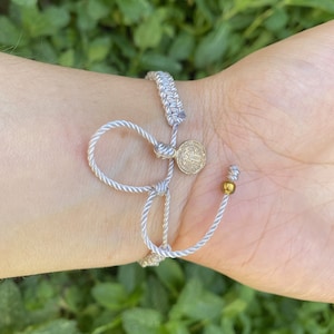 May include: A silver-gray bracelet with a woven band and adjustable cord. A small gold-colored coin charm and a gold bead accent the design. The bracelet is tied on the wrist.