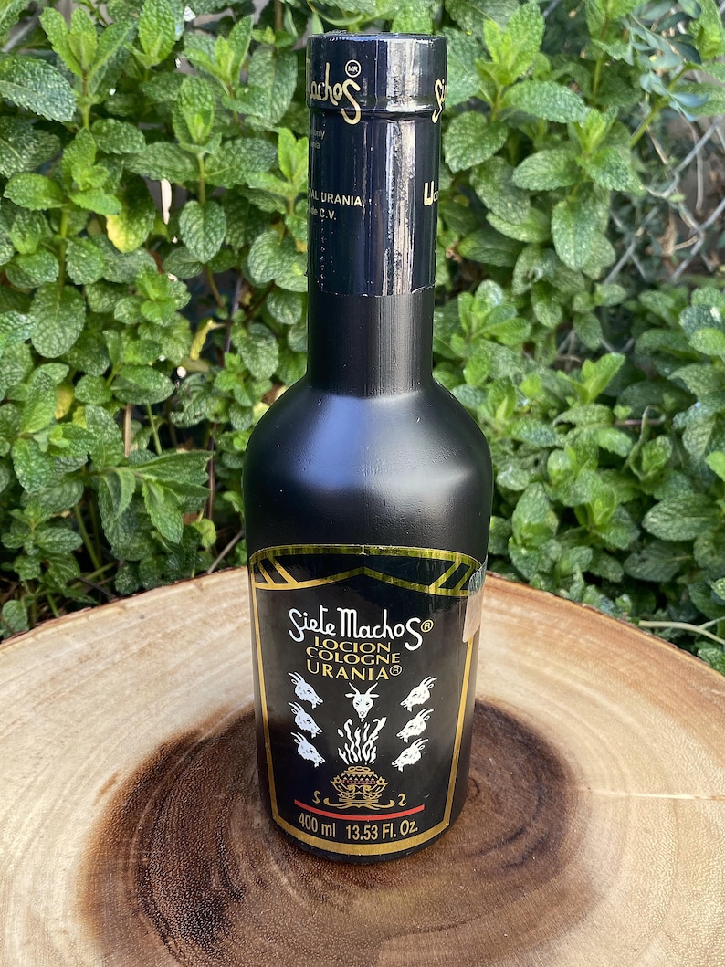 May include: A black cologne bottle with gold lettering and a label featuring the text "Siete Machos Locion Cologne Urania". The bottle contains 400 ml or 13.53 fl oz of cologne. The bottle is set against a green leafy background.