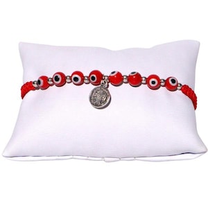 May include: Red beaded bracelet featuring evil eye beads and a silver charm. The bracelet is strung on a red cord and rests on a white pillow. The evil eye beads are red with a black and white eye design.