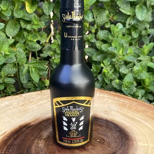 May include: A black cologne bottle with gold lettering and accents. The label reads "Siete Machos Locion Colonia Urania" and includes the volume: 110 ml (3.72 fl oz). The bottle is set against a background of green foliage.