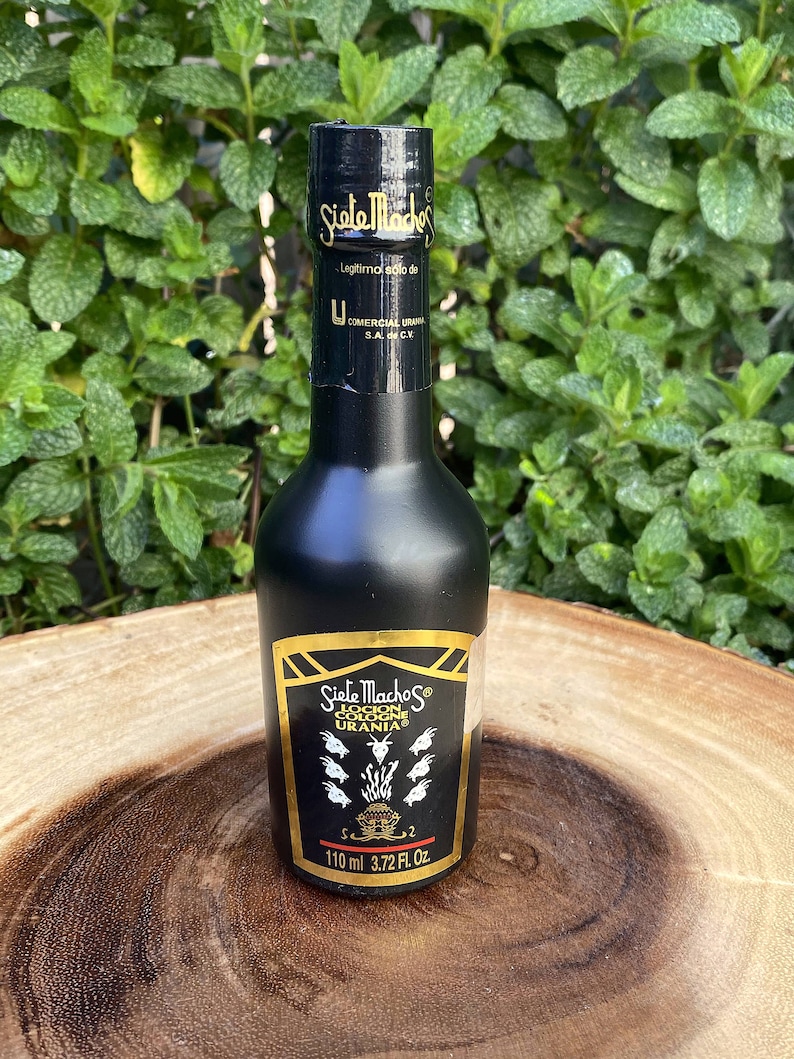 May include: A black cologne bottle with gold lettering and accents. The label reads "Siete Machos Locion Colonia Urania" and includes the volume: 110 ml (3.72 fl oz). The bottle is set against a background of green foliage.