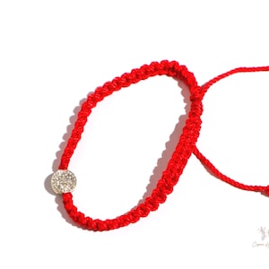 May include: A bright red woven bracelet with a silver-colored circular charm. The bracelet is adjustable with a sliding knot closure. The charm appears to be encrusted with small, clear stones. The bracelet is displayed on a white background.