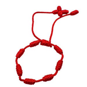 May include: A red knotted bracelet with a cross-shaped accent. The bracelet is made of a thick, woven red cord and features several large knots along its circular design. The cross is at the top, and the bracelet is against a white background.