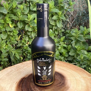 May include: A black cologne bottle with gold accents and the text "Siete Machos Locion Cologne Urania" is displayed on a wooden surface. The bottle contains 400 ml or 13.53 fl. oz. of cologne. The background features green foliage.