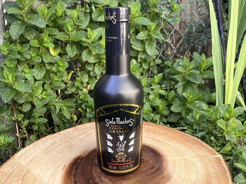 May include: A black cologne bottle with gold accents and the text "Siete Machos Locion Cologne Urania" is displayed on a wooden surface. The bottle contains 400 ml or 13.53 fl. oz. of cologne. The background features green foliage.