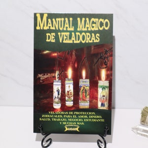 May include: A book titled "Manual Magico de Veladoras" with a green and gold cover, displayed on a black stand. The cover features images of lit candles and text in Spanish. Sage bundles and a crystal are in the foreground.