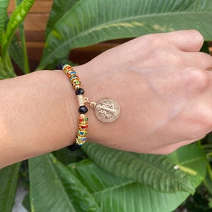 May include: A beaded bracelet with a gold charm. The bracelet features black, gold, and colorful beads. The gold charm has a detailed design. The bracelet is worn on a wrist.