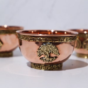 May include: Copper bowls with brass accents. Each bowl features a different embossed design: a tree, a hand, and a triple moon symbol. The bowls have a decorative band around the rim and base, and are displayed on a white surface.