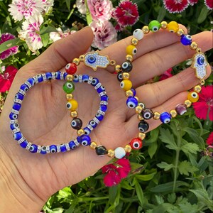 May include: Two beaded bracelets featuring colorful evil eye beads and gold accents. One bracelet is primarily blue with clear crystal spacers, while the other has a variety of colors and a gold Hamsa hand charm.