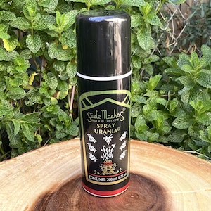 May include: A black spray bottle with white and gold accents, labeled "Siete Machos Locion Cologne Spray Urania." The bottle has a black cap and a red base. The background features green foliage and a wooden surface.