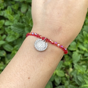 May include: Red beaded bracelet with a silver charm. The bracelet features small, faceted red beads and a round silver charm with a religious figure. The charm is encrusted with small, clear crystals.