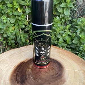 May include: A black spray bottle of "Siete Machos Spray Urania" cologne, with gold and white accents. The bottle is on a wooden surface, with a green leafy background. The bottle contains 6.75 fl oz (200 ml).