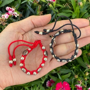 Könnte beinhalten: Zwei handgefertigte Armbänder, eines rot und eines schwarz, werden vor einem Hintergrund aus grünem Laub und Blumen gezeigt. Jedes Armband hat silberfarbene Perlen und dekorative Elemente mit verstellbaren Kordelverschlüssen.