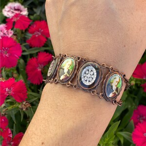 May include: A copper-toned bracelet featuring oval-shaped religious images. The bracelet has a chain-link design and is worn on a wrist. The images are framed with decorative metalwork. The background includes red flowers.