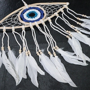 May include: A handmade dreamcatcher with an eye-shaped frame. The center features a blue and white woven design. White feathers hang from the bottom, and small wooden beads are interspersed along the hanging cords. The dreamcatcher is designed to be hung.