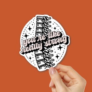 May include: A white sticker with a black illustration of a spine and the text "you're like really strong" in pink and black. The sticker is held by a hand against an orange background.
