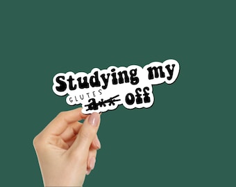 Puede incluir: Una pegatina blanca con texto negro que dice "Studying my glutes off". La pegatina está sujeta por una mano sobre un fondo verde oscuro. El texto está en una fuente retro en negrita.