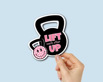 Puede incluir: Una pegatina negra con forma de pesa rusa con las palabras "LIFT each other UP" en rosa. La pesa rusa tiene una carita sonriente con un patrón de cuadros rosas y blancos. La pegatina está sujeta por una mano sobre un fondo azul claro.