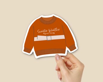 Puede incluir: Una pegatina naranja con un diseño de suéter de dibujos animados. El suéter tiene una franja blanca en el pecho con las palabras "Sweater Weather Physical Therapy" en una fuente de escritura. La pegatina está sujeta por una mano.