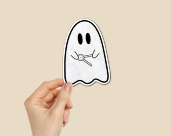 Puede incluir: Una pegatina de fantasma blanco con un contorno negro y dos ojos ovalados. El fantasma sostiene una llave inglesa. La pegatina está sujeta por una mano sobre un fondo beige.