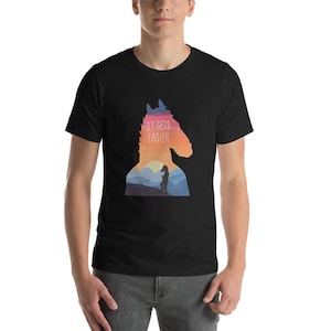 Puede incluir: Una camiseta negra con la silueta de un caballo y una figura caminando en un paisaje. El texto "IT GETS EASIER" está impreso en la camiseta.