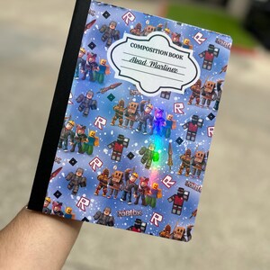 May include: A composition book with a blue cover featuring a pattern of Roblox characters and the letter "R". The cover includes the text "COMPOSITION BOOK" and a label with the name "Abad Martinez".