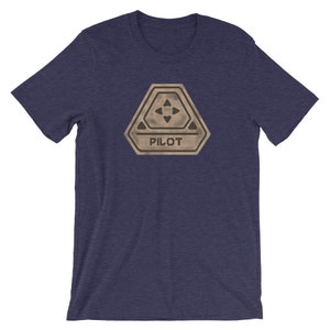 May include: A navy blue t-shirt with a brown and black graphic of a pilot badge. The badge has a five-pointed star with four arrows pointing inward and the word "PILOT" printed below.