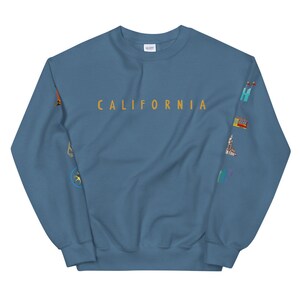 Puede incluir: Una sudadera azul con la palabra "CALIFORNIA" impresa en dorado en la parte delantera. Las mangas presentan una variedad de iconos coloridos con temática californiana, incluyendo una palmera, una tabla de surf y un tranvía.