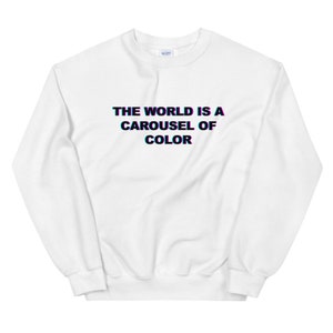 Puede incluir: Sudadera blanca con el texto "THE WORLD IS A CAROUSEL OF COLOR" en negro con un contorno azul y rojo. La sudadera tiene cuello redondo y mangas largas.
