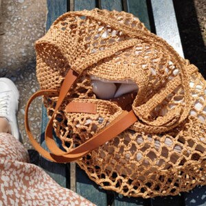 May include: A tan crocheted mesh bag with brown leather straps. The bag is sitting on a wooden bench.