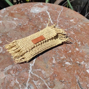 May include: A beige crocheted clutch with a brown leather tag that says "Nella" and fringe details.