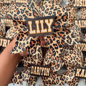 Op de afbeelding: Een verzameling haarstrikken met een beige, zwart en oranje luipaardprintpatroon met verspreide doorzichtige strass-steentjes. Elke strik heeft een zwart rechthoekig middenstuk met een naam in wit en oranje, waaronder LILY en DARIYON.