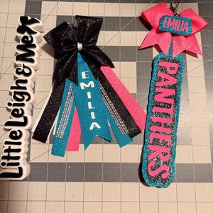 May include: Two cheer bows with attached ribbons and a white 3D sign reading Little Leigh & Me. One bow is black with teal ribbons featuring the name EMILIA and rhinestone trim. The other is pink with a teal tag reading EMILIA and PANTHERS.