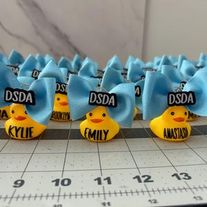 May include: A collection of yellow rubber duck keychains with light blue bows. Each duck has a name printed on it, and a black and white label that says "DSDA". The ducks are arranged on a grid.