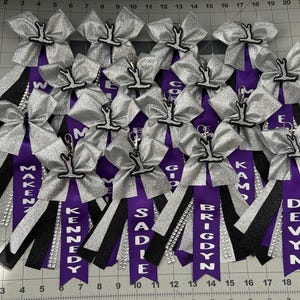 Może przedstawiać: Kolekcja kokardek dla cheerleaderek ze srebrnymi kokardami, czarnymi i srebrnymi wstążkami oraz fioletowymi wstążkami z imionami takimi jak 'Makena' i 'Kennedy'. Każda kokarda ma zawieszkę z figurką cheerleadera.